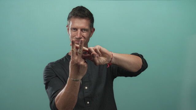 Man holding up two fingers with hands forward wearing casual black shirt and visible bracelet in studio with teal wall; playful.