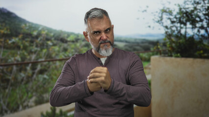 Man with clasped hands and furrowed brow glaring while leaning forward on a terrace in a forest setting near green foliage; anger defiance.