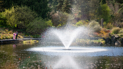 A vibrant park scene featuring a powerful water fountain spraying high into a tranquil pond, observed by an elegantly dressed couple on a sunny day.