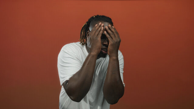 Young african american man covering his face with both hands in a studio wearing a white t shirt against an orange backdrop; anxiety. - Powered by Adobe