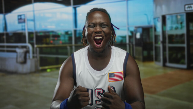 Young man smiling with clenched fists, wearing usa basketball jersey with flag patch and blue wristbands, dreadlocks visible, standing at airport terminal entrance; pride celebration. - Powered by Adobe