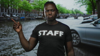Man points finger on street beside amsterdam canal wearing staff t shirt, stern expression and raised arm; confidence duty.