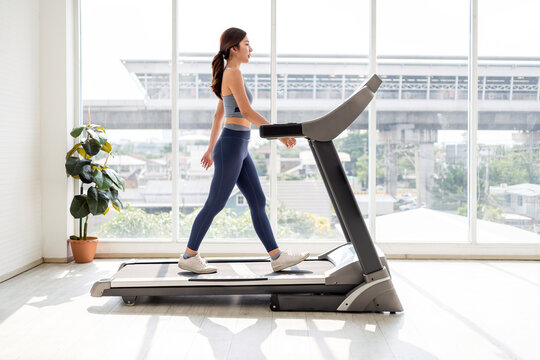 A young Asian woman in activewear walking on a treadmill in a bright home gym with large windows and urban view. New year resolution, treat yourself.