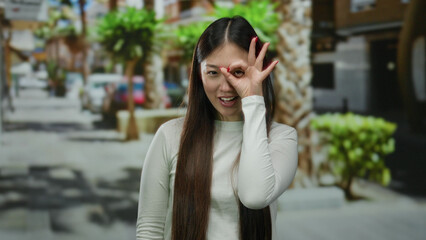 Woman making playful gesture on city street surrounded by urban greenery during daylight, expressing joy and confidence.
