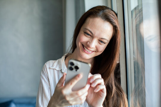 Adult woman smiling at smartphone by window at home