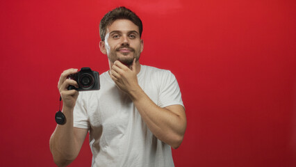 Naklejka premium Man photographer holding camera and gesturing with hand to chin in studio against red wall; thoughtful introspection.