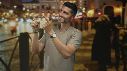 Man points both index fingers upward on a busy city street at night, smiling and squinting; playful confidence.