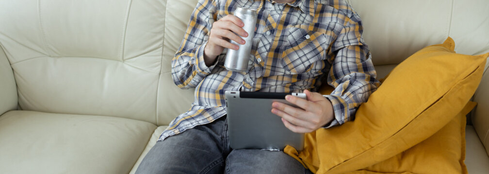 Man relaxing on sofa with tablet and aluminum drink can in hand. Casual leisure, digital lifestyle, and technology use in everyday home environment.