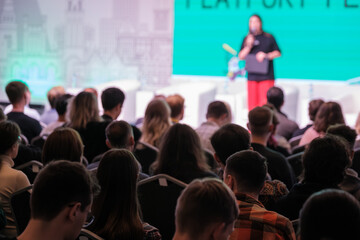 Fototapeta premium Crowd attends conference listening to speaker on stage with large blue backdrop and bright screen