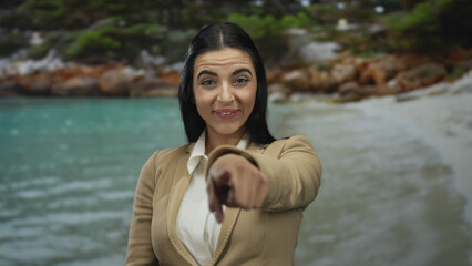 Woman gesturing in laughter at a seaside location wearing a blazer, with a background of turquoise sea and sandy beach, conveying a joyful and relaxed atmosphere.