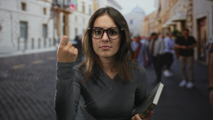 Woman wearing glasses holding book beckons with fingers toward camera in a crowded city street; defiance challenge.