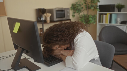 Hispanic woman resting head on computer keyboard at work desk in office late after long shift;...