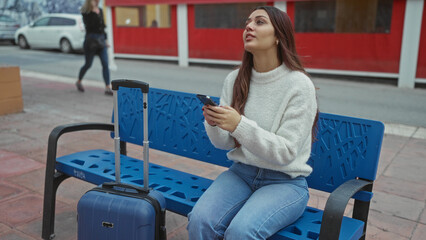 Woman sitting on a street bench using a smartphone with a blue suitcase nearby, capturing an urban travel scene with casual attire on a sidewalk outdoors.