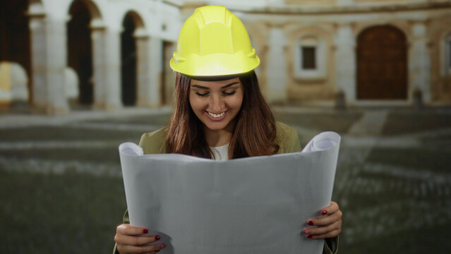 Hispanic female architect smiles while reviewing blueprints at an old university exterior, wearing a yellow hard hat.