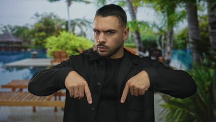 Young hispanic man pointing both index fingers down at poolside building walkway outdoors; assertion.