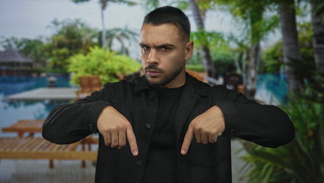 Young hispanic man pointing both index fingers down at poolside building walkway outdoors; assertion. - Powered by Adobe