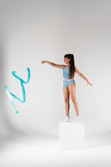 A focused young rhythmic gymnast with a ponytail, wearing a blue and white patterned leotard, gracefully performs with a light blue ribbon while balancing on a white cube in a studio