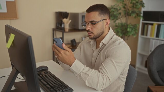 Man holds smartphone at computer desk with monitor in building during work hours; productivity efficiency focus.