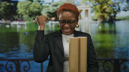 Young woman wearing glasses holds rolled blueprints confidently and pumps fist near building; success victory.