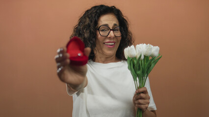 Middle-aged hispanic woman holding engagement ring and flowers against brown wall, showcasing joyful proposal moment.
