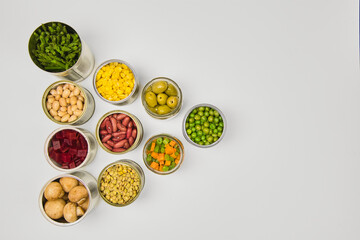 Open cans of beans, mushrooms, carrots, corn, peas, green beans, lentils, asparagus, and olives on a white background with copy space.