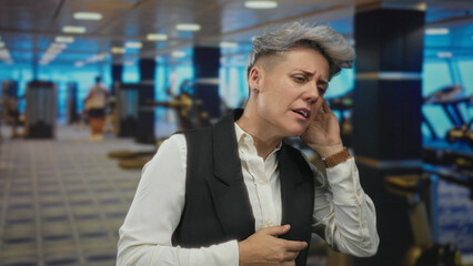 Woman with short hair clutches chest while raising hand by face in indoor gymnasium; concern healing support.