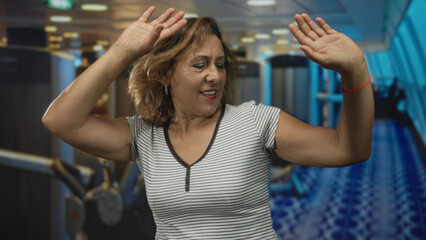 Woman waving hands inside a gym training area with exercise machines visible in a modern building; happiness celebration.
