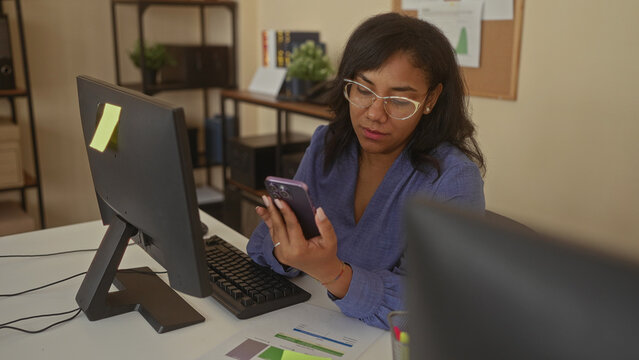 Woman reading smartphone screen with one hand at desk next to computer monitor in office building; concentration.