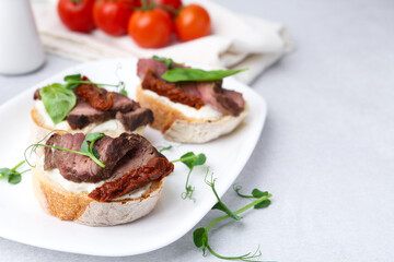 Bruschettas with roast beef, cream cheese, basil, sun-dried tomatoes and fresh vegetables on light grey table, closeup. Space for text