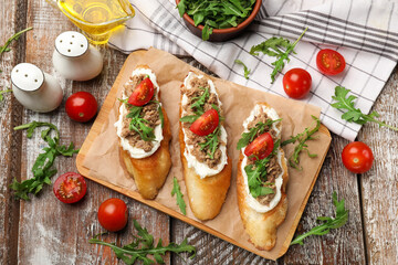 Delicious bruschettas with tuna, tomatoes, oil and arugula on wooden table, flat lay