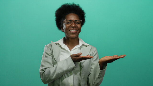 Woman smiling with hands extended in presentation over an isolated green background wearing glasses and suit offering a friendly gesture indicating welcome or introduction