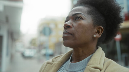 Woman standing on city street in urban environment wearing coat looking thoughtful with afro hairstyle outdoors on a cloudy day showcasing independence and confidence.