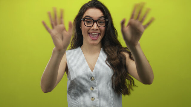 Woman with glasses wearing striped vest waves hands and smiles against green studio wall; warm friendly greeting positivity. - Powered by Adobe