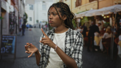 Woman points finger at ring on her hand on a cobblestone street terrace outside restaurant wearing plaid shirt and bracelet; mild concern.