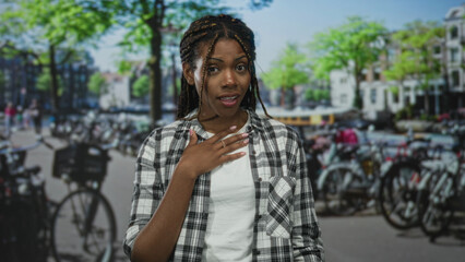 African american woman with braided hair hand on chest looking surprised on amsterdam street lined with bicycles; surprise apology.