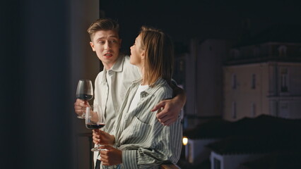 Relaxed couple talking balcony at night. Smiling man embracing girl holding wine