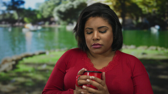 Woman in red top holding coffee cup in park setting, suggesting a reflective moment outdoors. - Powered by Adobe