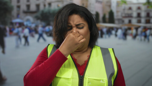 Young woman in a reflective vest sneezing on a bustling city street showcasing a diverse urban environment.