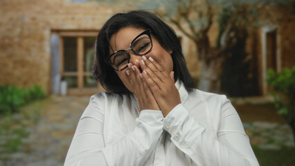 Woman smiling wearing glasses with hands over mouth in outdoor city setting featuring rustic stone...