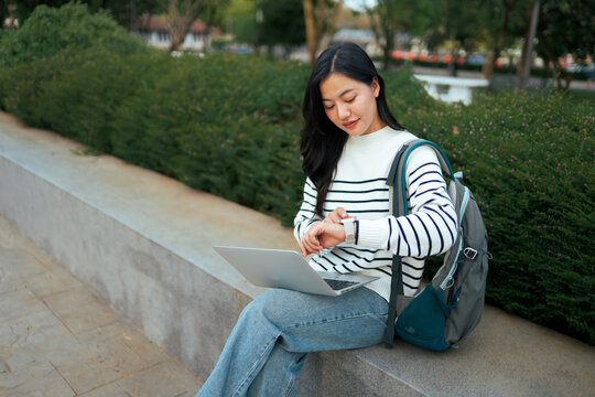 Asian student checking smartwatch while using laptop outdoors