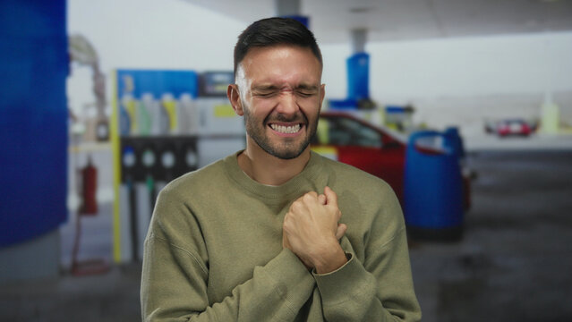 Young man at a gas station outdoors clutches his chest in pain, expressing discomfort, with blurred background of pumps and a red car, highlighting an emotional moment. - Powered by Adobe
