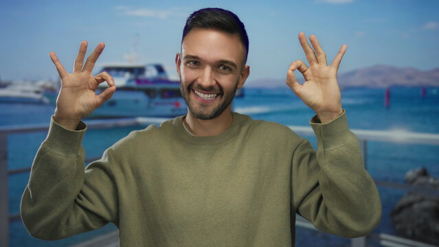 Young man making ok gesture with both hands and smiling on a seaside promenade with boats in the background on a sunny day
