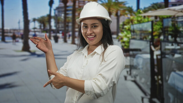 Woman in white hardhat clapping hands on street outside restaurant terrace while reviewing plans; confidence design planning.