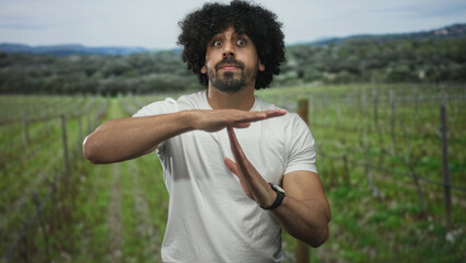 Obraz premium Man with curly hair and beard makes timeout sign with both hands in a forest among rows of vines, wearing a white t shirt and looking at camera; determination.