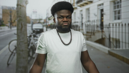 Fototapeta premium Man frowning and looking sideways wearing beaded necklace on a street with rowhouses and parked bicycles; urban resilience frustration.