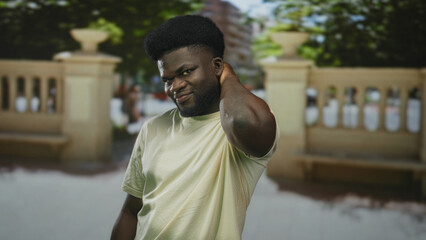 Fototapeta premium Man touching neck with right hand on street near stone balustrade, wearing light t shirt and slight smile; playful selfconscious.