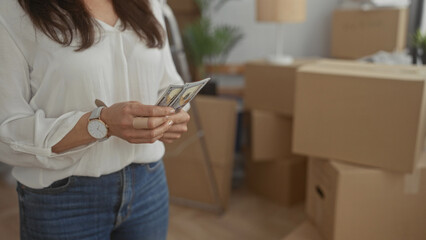 Woman counting dollars in new home with moving boxes in background suggesting recent relocation and...
