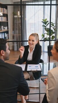 Vertical shot of a smiling businesswoman or HR manager conducting an interview or a consulting session with two applicants or clients across a glass table in a modern office meeting room.