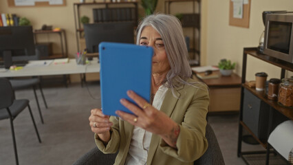Senior woman with grey hair takes a selfie using a tablet in a modern office setting, capturing a moment of leisure at work in a comfortable indoor environment.