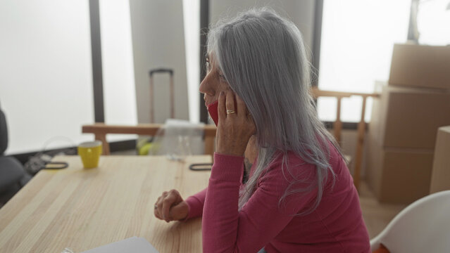 Woman talking on phone in modern living room with grey hair wearing pink sweater surrounded by moving boxes offering a sense of new beginnings and home transition.